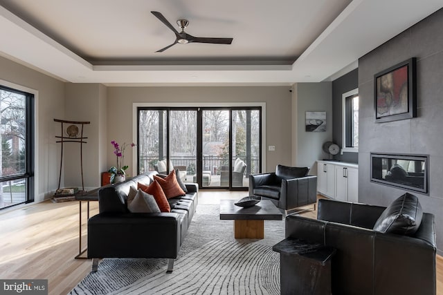 living room featuring a tile fireplace, ceiling fan, light hardwood / wood-style floors, and a tray ceiling