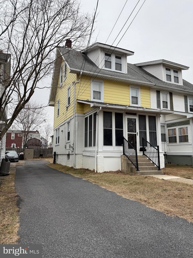 american foursquare style home featuring a chimney, aphalt driveway, entry steps, a sunroom, and roof with shingles