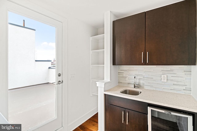 bar with wine cooler, dark brown cabinetry, sink, light stone countertops, and backsplash