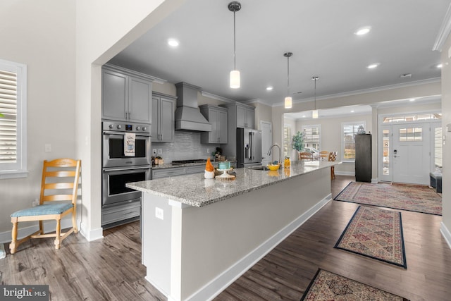 kitchen featuring appliances with stainless steel finishes, gray cabinets, custom range hood, and a center island with sink