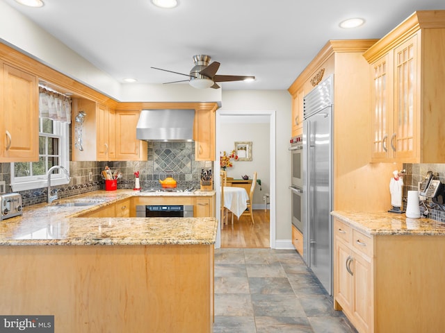 kitchen with light brown cabinetry, sink, kitchen peninsula, stainless steel appliances, and wall chimney exhaust hood