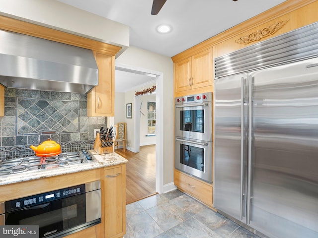 kitchen featuring appliances with stainless steel finishes, backsplash, range hood, light stone counters, and light brown cabinetry