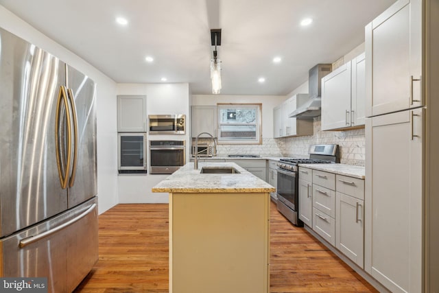 kitchen with appliances with stainless steel finishes, decorative light fixtures, a kitchen island with sink, wall chimney range hood, and light hardwood / wood-style flooring