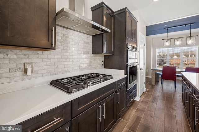 kitchen featuring dark brown cabinets, hanging light fixtures, stainless steel appliances, decorative backsplash, and wall chimney range hood