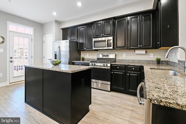 kitchen featuring light wood finished floors, a kitchen island, dark cabinets, stainless steel appliances, and a sink