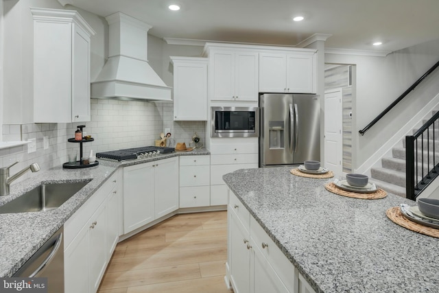 kitchen with white cabinetry, sink, light stone counters, stainless steel appliances, and custom range hood