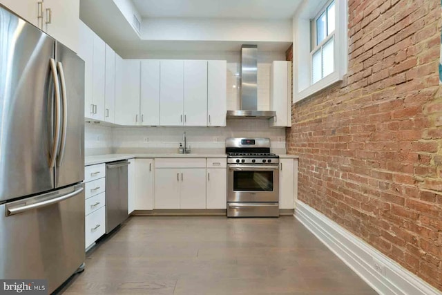 kitchen featuring brick wall, appliances with stainless steel finishes, white cabinetry, sink, and wall chimney range hood