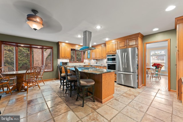 kitchen with a breakfast bar, a center island, dark stone counters, island exhaust hood, and stainless steel appliances