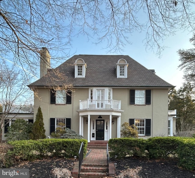 colonial inspired home with a balcony, a chimney, and stucco siding