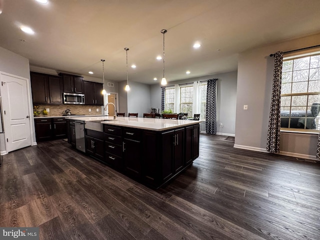 kitchen featuring dark wood finished floors, stainless steel appliances, light countertops, backsplash, and an island with sink