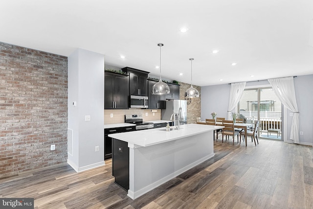 kitchen featuring appliances with stainless steel finishes, dark cabinets, a kitchen island with sink, light countertops, and pendant lighting