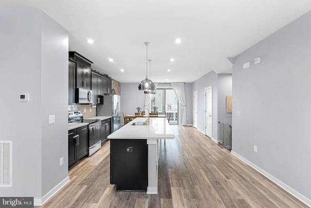 kitchen with a kitchen island with sink, stainless steel appliances, a sink, light countertops, and dark cabinetry