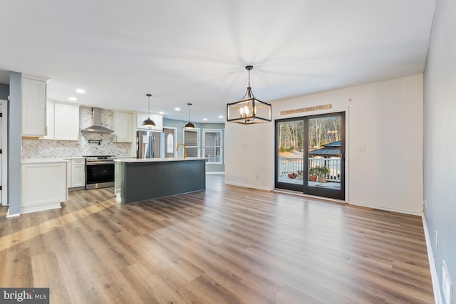 kitchen featuring wall chimney exhaust hood, appliances with stainless steel finishes, pendant lighting, a kitchen island with sink, and white cabinets