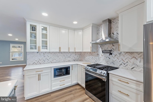 kitchen with wall chimney range hood, light wood-type flooring, white cabinets, and appliances with stainless steel finishes