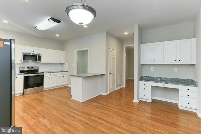 kitchen with appliances with stainless steel finishes, built in desk, white cabinets, dark stone counters, and light hardwood / wood-style flooring