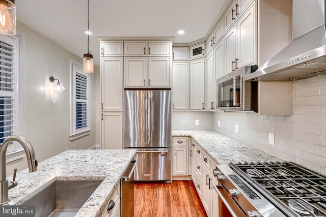 kitchen with light wood finished floors, wall chimney exhaust hood, hanging light fixtures, stainless steel appliances, and a sink