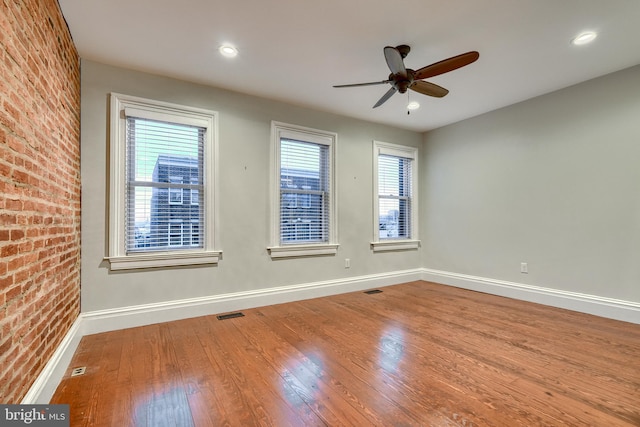 spare room with ceiling fan, a wealth of natural light, wood-type flooring, and brick wall