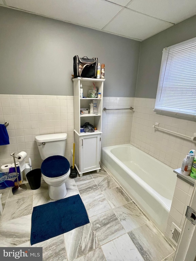 bathroom featuring a paneled ceiling, toilet, a bathtub, and tile walls