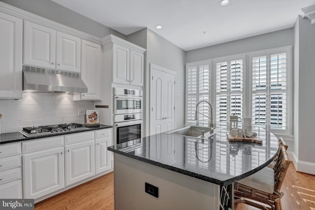 kitchen with sink, appliances with stainless steel finishes, dark stone countertops, white cabinets, and a center island with sink