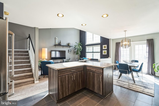 kitchen with dark brown cabinetry, a chandelier, stainless steel refrigerator, a kitchen island, and light stone countertops