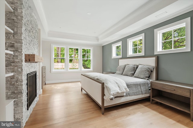 bedroom with a stone fireplace, light wood-style flooring, baseboards, ornamental molding, and a raised ceiling