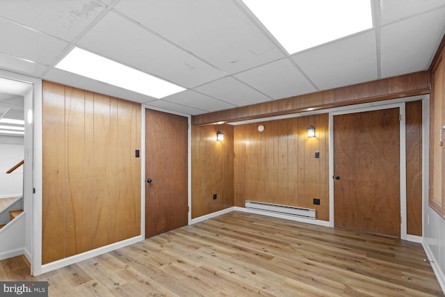 basement featuring a baseboard radiator, wooden walls, light hardwood / wood-style flooring, and a drop ceiling