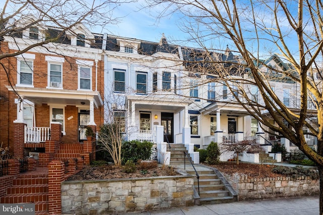 townhome / multi-family property featuring mansard roof, cooling unit, covered porch, brick siding, and stairs