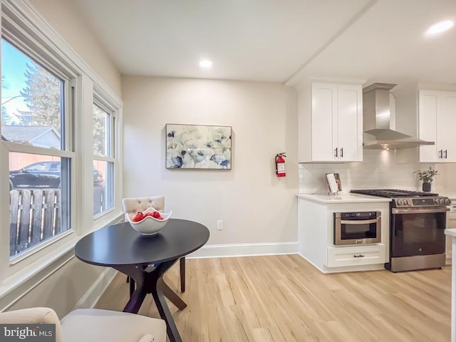 kitchen with appliances with stainless steel finishes, tasteful backsplash, white cabinets, wall chimney range hood, and light hardwood / wood-style flooring