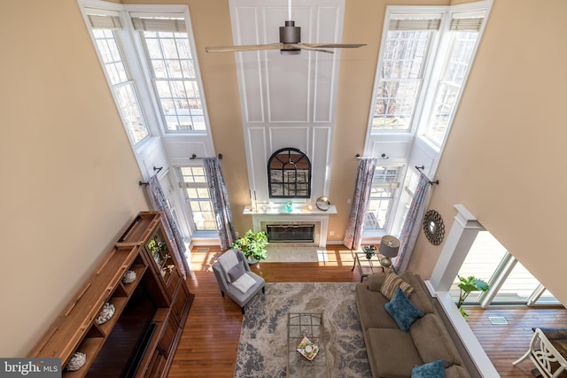 living room with a glass covered fireplace, ceiling fan, a towering ceiling, and wood finished floors