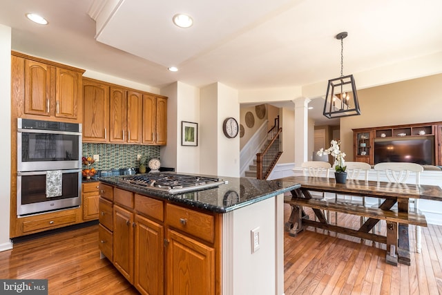 kitchen with light wood-type flooring, tasteful backsplash, stainless steel appliances, brown cabinetry, and ornate columns