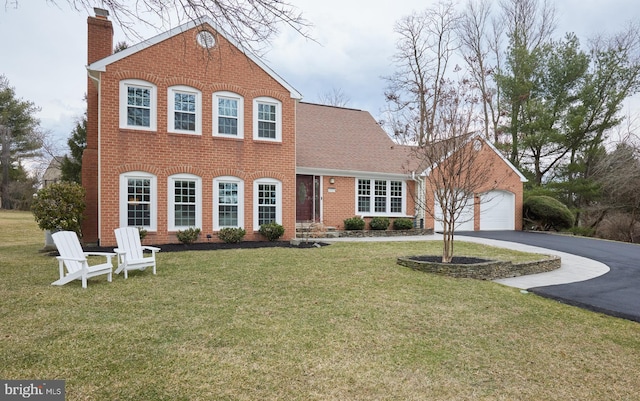 colonial inspired home featuring aphalt driveway, brick siding, a chimney, a shingled roof, and a front lawn