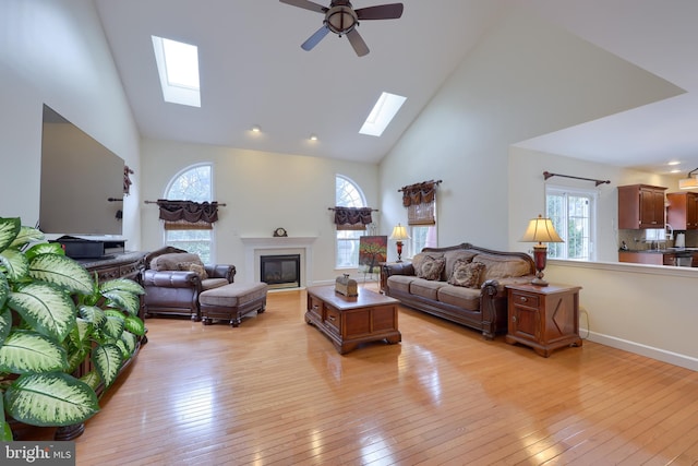 living room featuring high vaulted ceiling, light wood finished floors, a skylight, and a glass covered fireplace