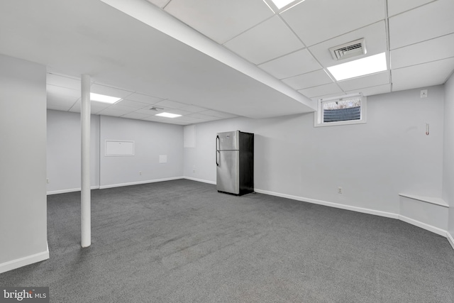 finished below grade area featuring a paneled ceiling, visible vents, baseboards, freestanding refrigerator, and dark colored carpet