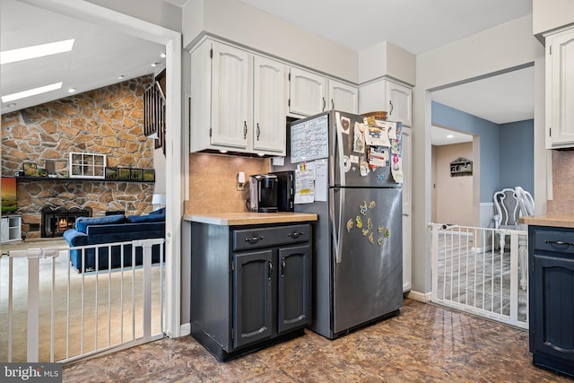 kitchen featuring lofted ceiling with skylight, a stone fireplace, white cabinetry, stainless steel fridge, and backsplash