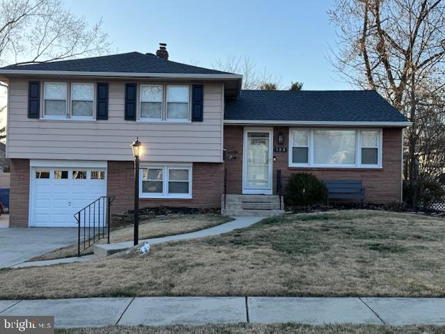 split level home with concrete driveway, a front yard, an attached garage, brick siding, and a chimney