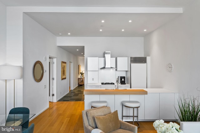 kitchen with white cabinets, a kitchen bar, kitchen peninsula, white built in fridge, and wall chimney range hood