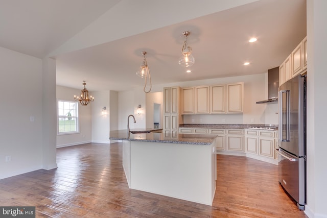 kitchen with pendant lighting, wall chimney range hood, stainless steel appliances, light hardwood / wood-style floors, and an island with sink