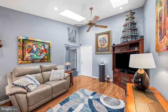 living room with ceiling fan, a towering ceiling, light hardwood / wood-style floors, and a skylight