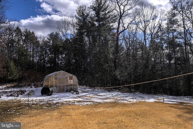 snow covered structure featuring a greenhouse, a wooded view, and an outdoor structure