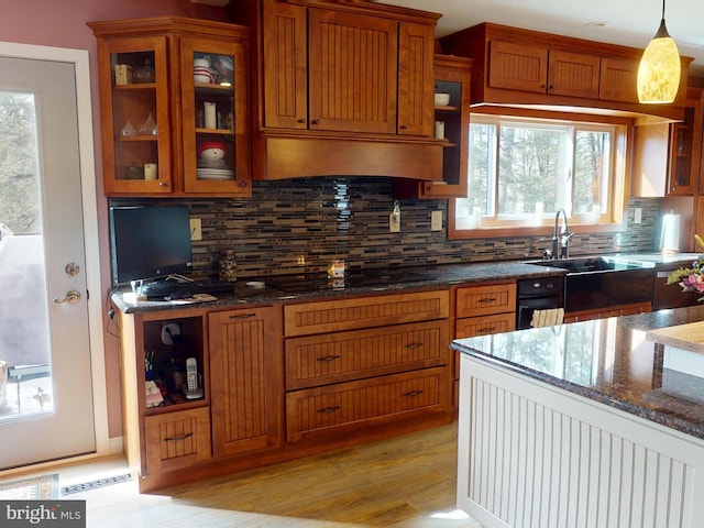 kitchen with range hood, black stovetop, brown cabinetry, glass insert cabinets, and dark stone counters