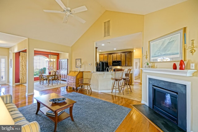 living area featuring light wood finished floors, baseboards, visible vents, a glass covered fireplace, and high vaulted ceiling