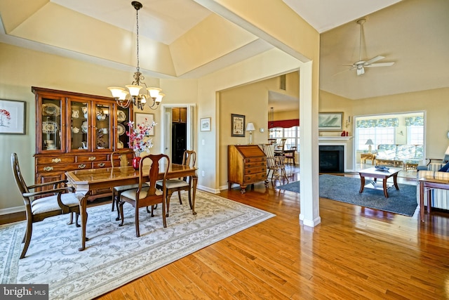 dining space with a fireplace, light wood finished floors, a raised ceiling, baseboards, and ceiling fan with notable chandelier