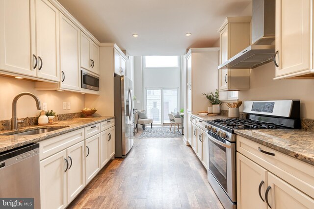 kitchen with stainless steel appliances, a sink, wall chimney range hood, light wood-type flooring, and light stone countertops