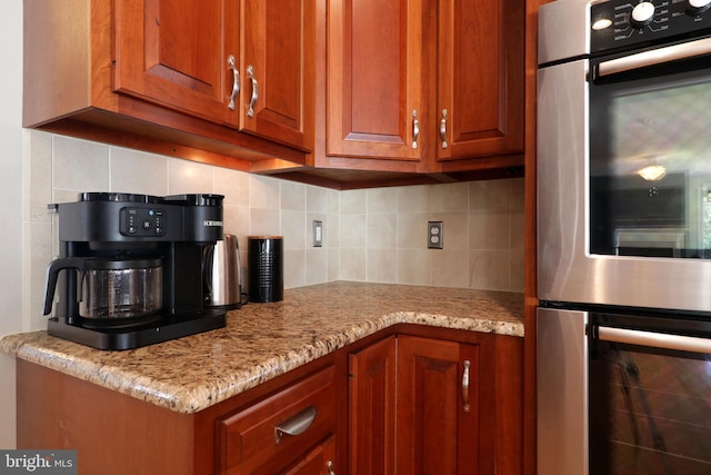 kitchen with light stone counters, stainless steel double oven, backsplash, and brown cabinetry