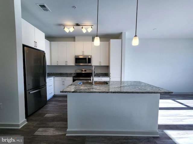 kitchen with white cabinetry, decorative light fixtures, dark stone counters, stainless steel appliances, and a kitchen island with sink