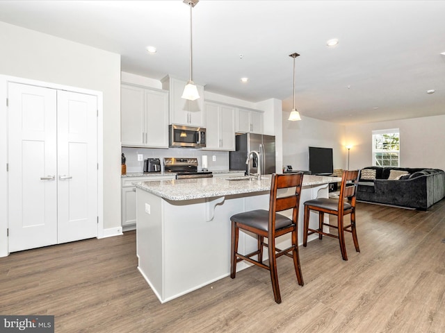 kitchen featuring appliances with stainless steel finishes, an island with sink, white cabinets, hanging light fixtures, and light stone countertops