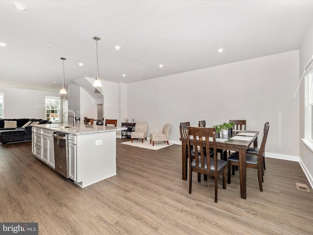 kitchen with dishwasher, a kitchen island with sink, hanging light fixtures, wood-type flooring, and light stone countertops