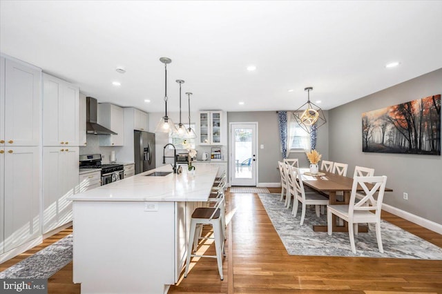 kitchen with a breakfast bar, stainless steel appliances, glass insert cabinets, a kitchen island with sink, and wall chimney range hood