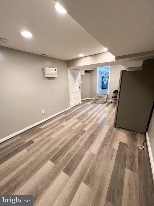 finished below grade area featuring a wall mounted air conditioner, baseboards, light wood-type flooring, and freestanding refrigerator