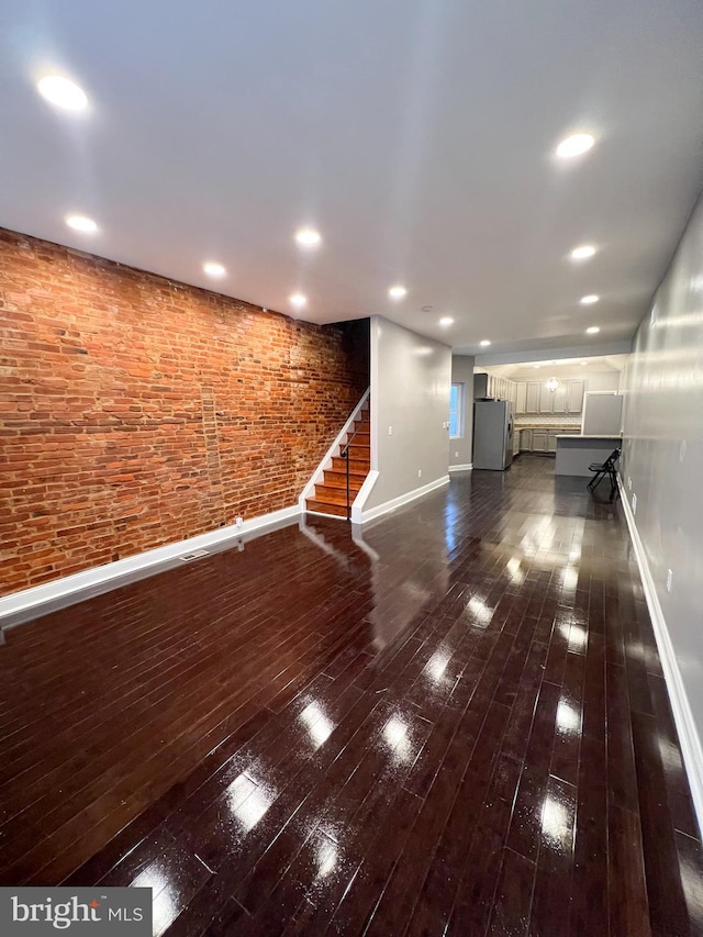 unfurnished living room featuring brick wall, baseboards, stairs, recessed lighting, and dark wood-style floors